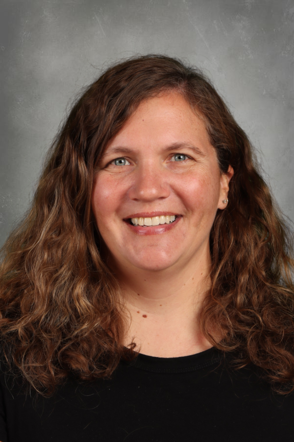A woman with curly brown hair, blue eyes, and wearing a black top, smiling against a gray background.