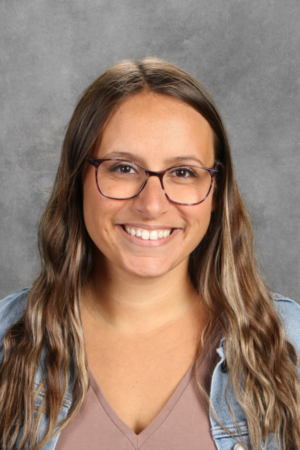 A young woman with long wavy hair, glasses, and a bright smile, wearing a beige top and a denim jacket, standing against a gray background.