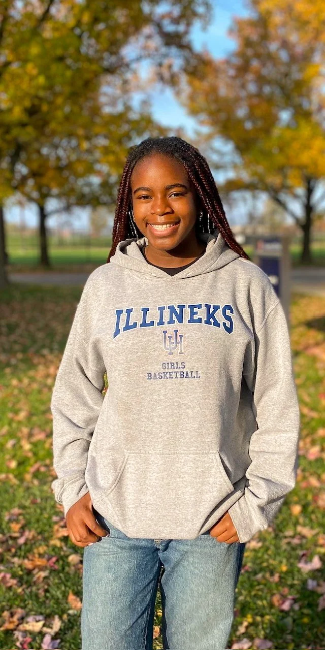 Smiling young girl in a gray hoodie with 'ILLINEKS GIRLS BASKETBALL' in blue text, standing outdoors on fall day with colorful trees and fallen leaves.