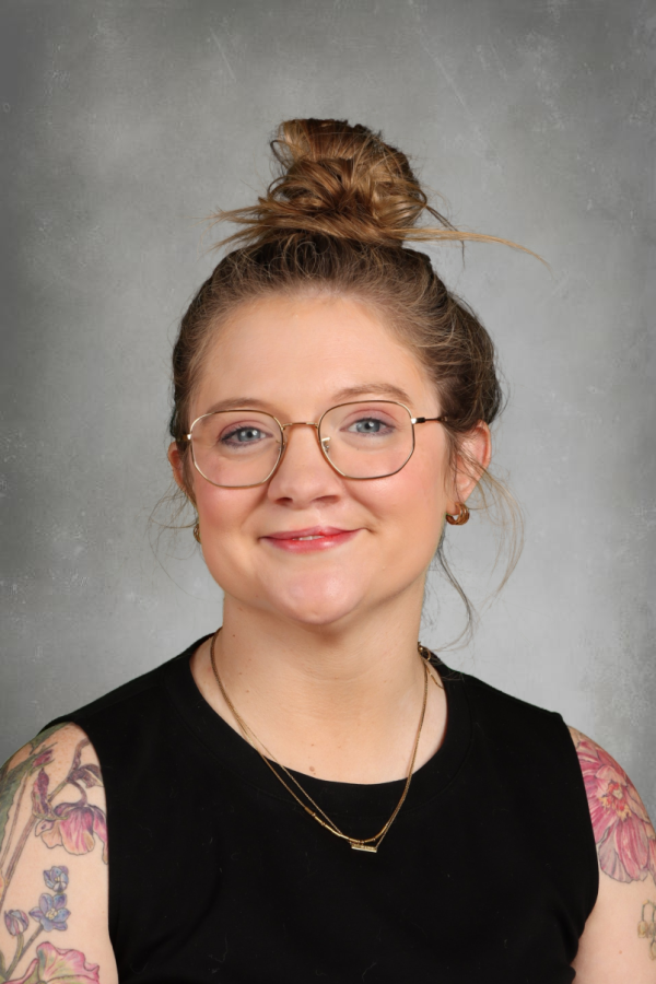 A young woman with glasses and tattoos smiling at the camera against a gray background.