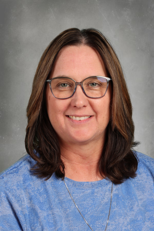 A woman with shoulder-length brown hair, wearing glasses and a blue top, smiling at the camera against a gray background.