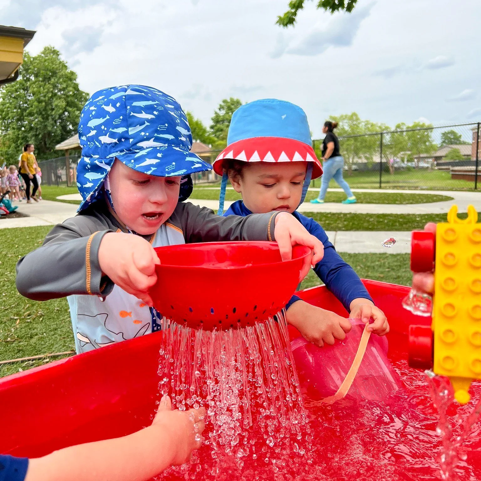 Two young children wearing bucket hats and raincoats playing with water in red plastic water tables in a park.