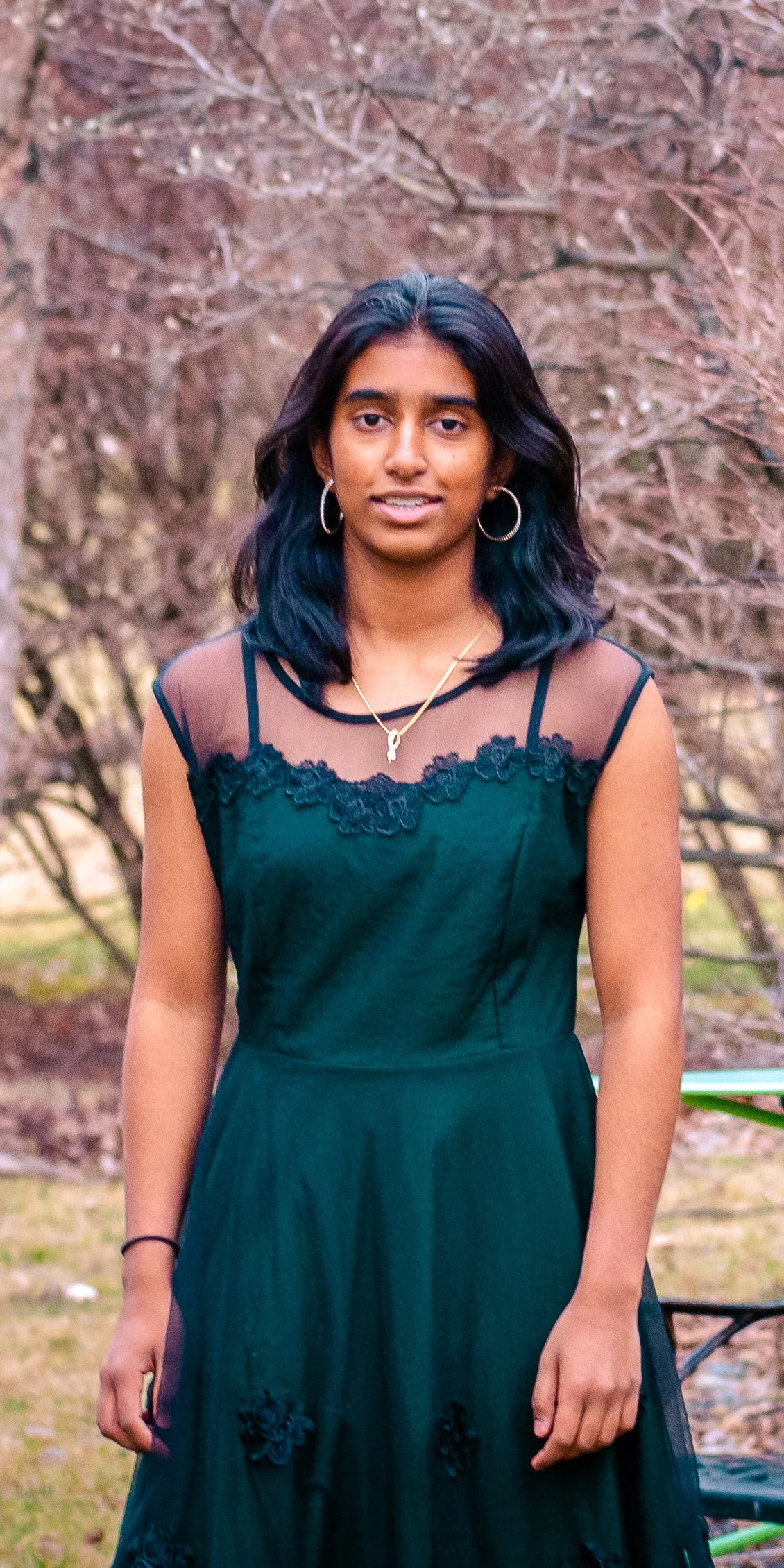 A young woman with dark hair wearing a black dress with lace details, standing outdoors with trees in the background.