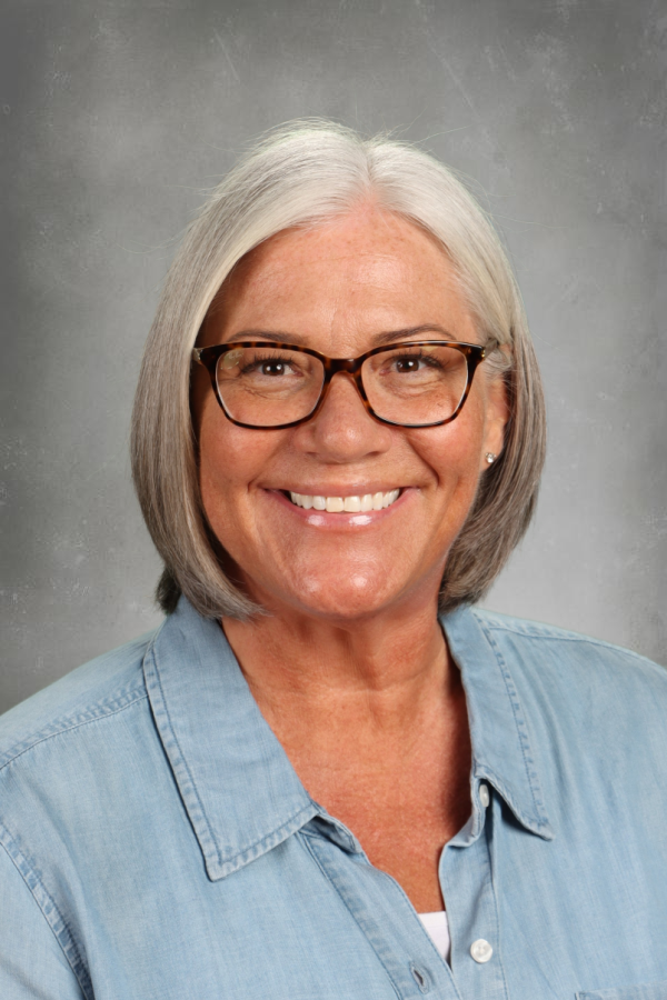 A smiling middle-aged woman with shoulder-length gray hair, wearing glasses and a light blue denim shirt against a gray background.
