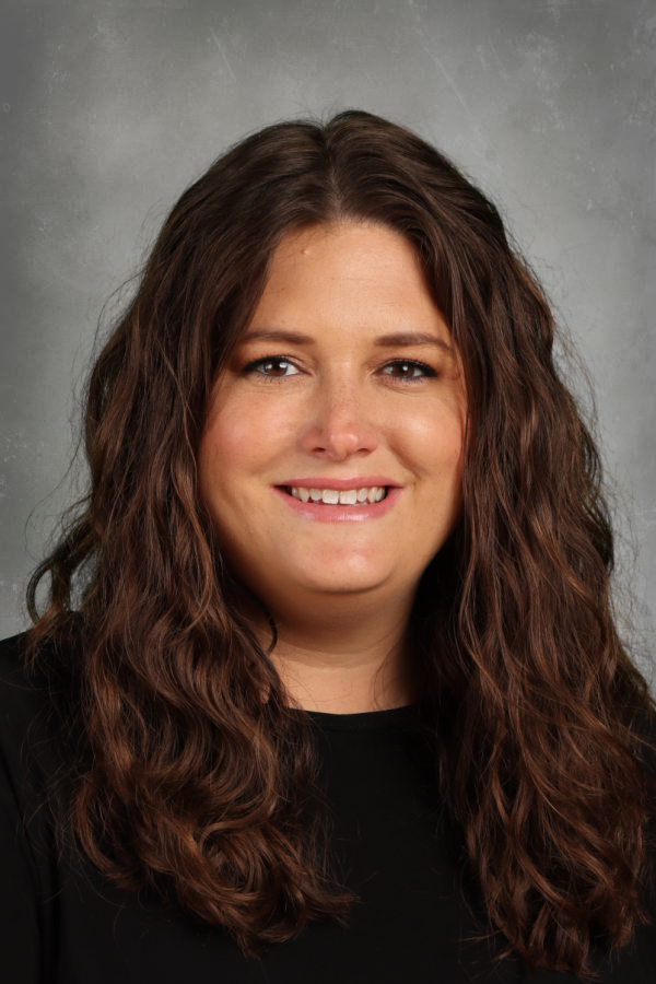 A woman with long, wavy brown hair smiling at the camera, wearing a black top with a gray background.
