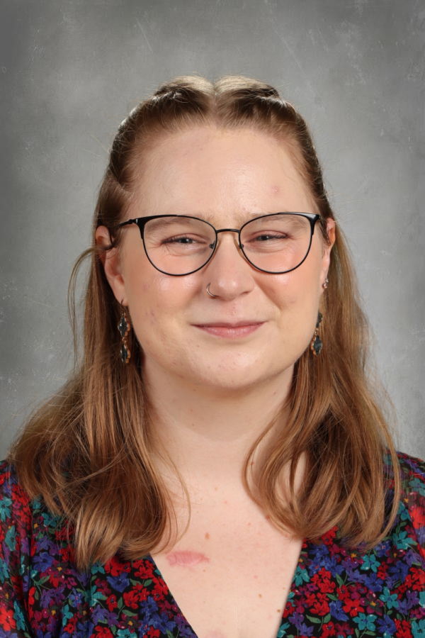 A young woman with long, light brown hair, wearing glasses, earrings, a nose ring, and a colorful floral top, smiling slightly against a gray background.
