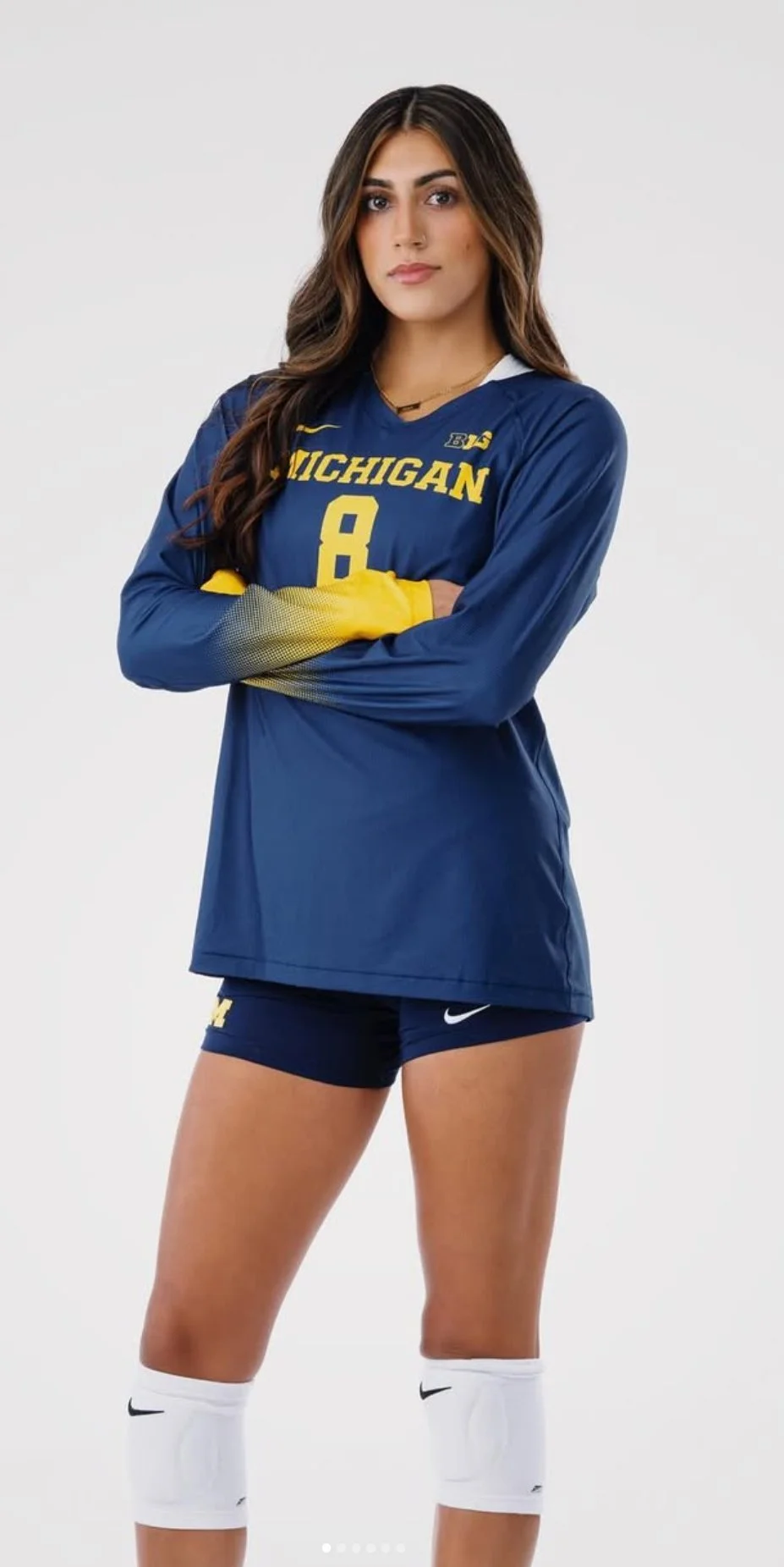 A female athlete in a blue Michigan volleyball uniform with yellow accents, standing with arms crossed and looking confident.