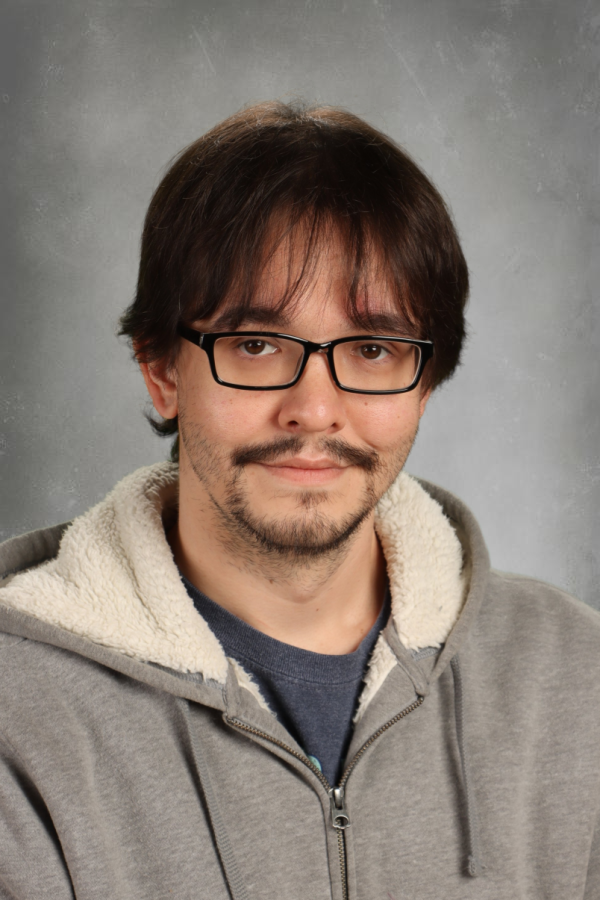 A man with glasses, dark brown hair, and a slight beard wearing a gray hoodie with a cream-colored fleece lining, standing against a gray background.