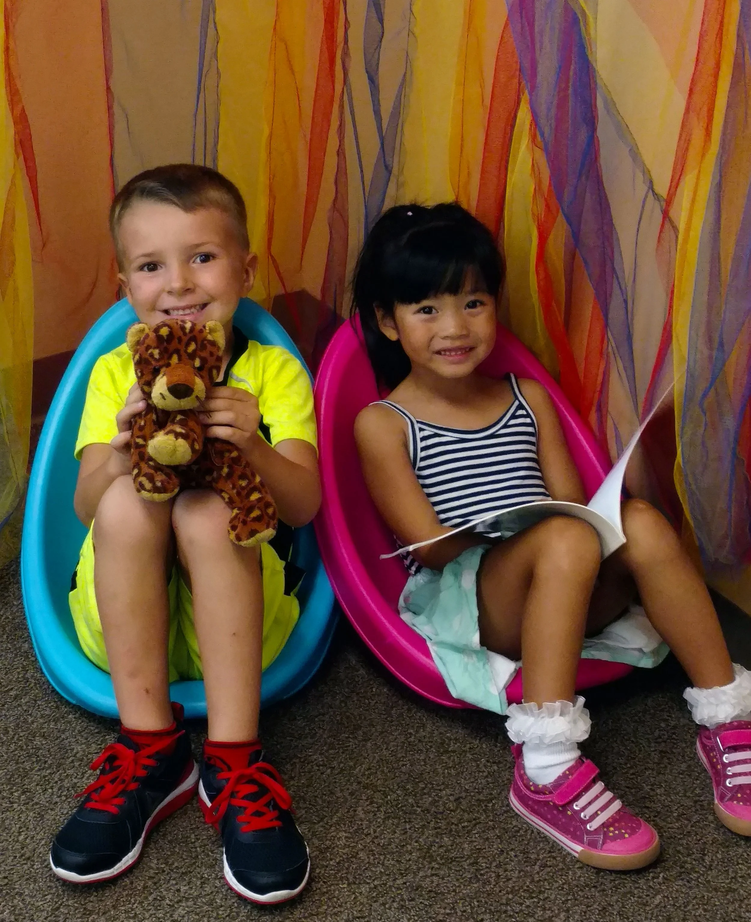 Two children sitting in colorful chairs against a decorative background. The boy on the left is smiling while holding a stuffed animal, a leopard. The girl on the right is smiling with a book in her lap.