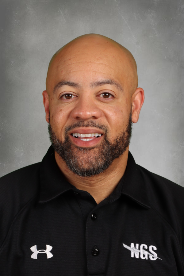 A man with a bald head and beard wearing a black polo shirt with 'NCS' and Under Armour logos, smiling in front of a gray background.