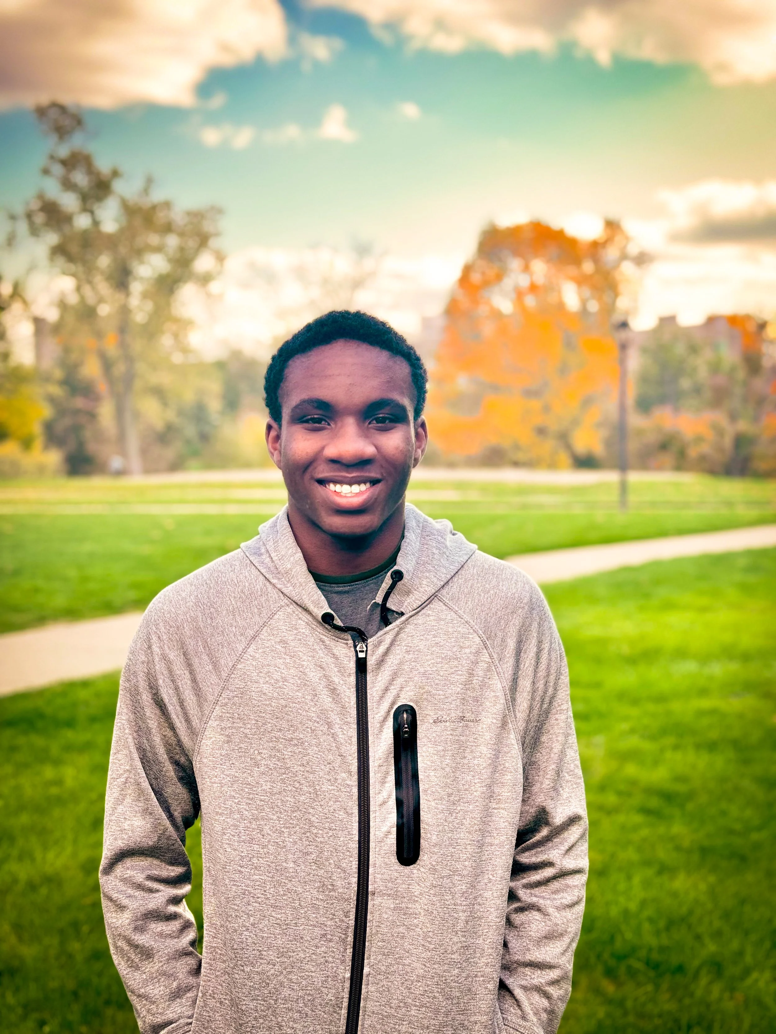 A smiling young man standing outdoors in a park during autumn, wearing a light-colored zip-up hoodie with a black zipper and pockets, with trees displaying fall foliage in the background.