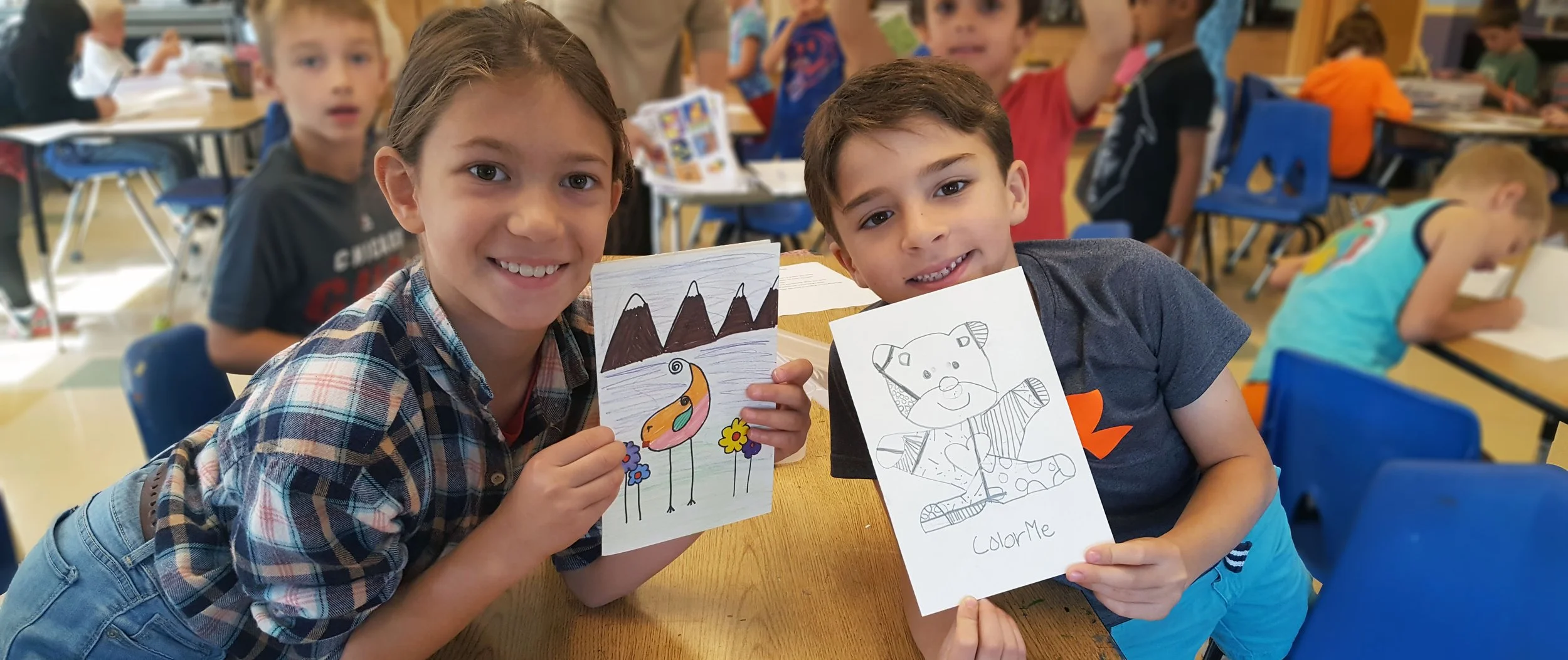 Two school children, a girl and a boy, sit at a table in a classroom, holding up their coloring pages and smiling at the camera. The girl has brown hair and wears a plaid shirt, while the boy has short brown hair and wears a dark T-shirt. In the background, other children are engaged in activities at their tables.