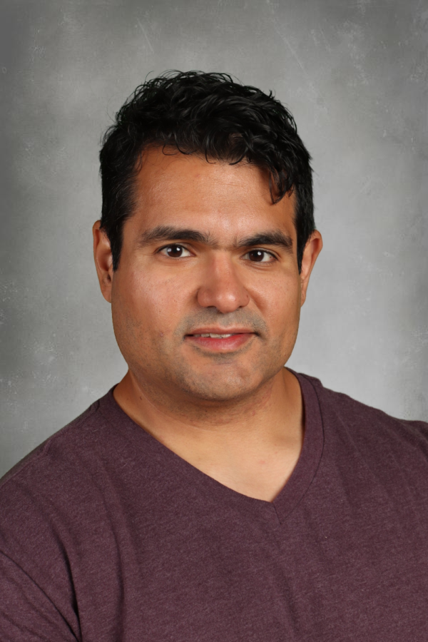 Portrait of a man with dark hair, wearing a maroon V-neck shirt, against a gray background.