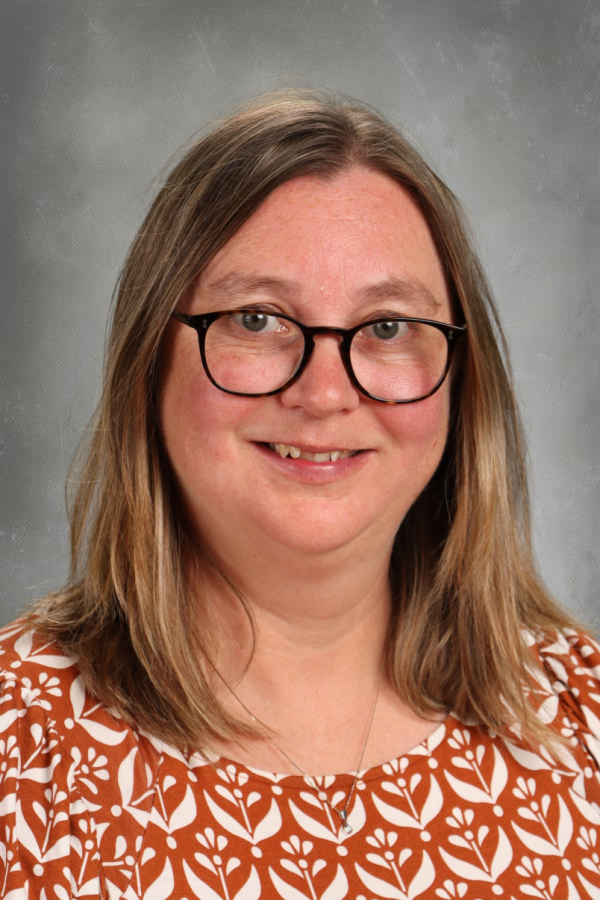 A woman with shoulder-length light brown hair, glasses, and a warm smile, wearing a brown and white patterned top against a plain gray background.