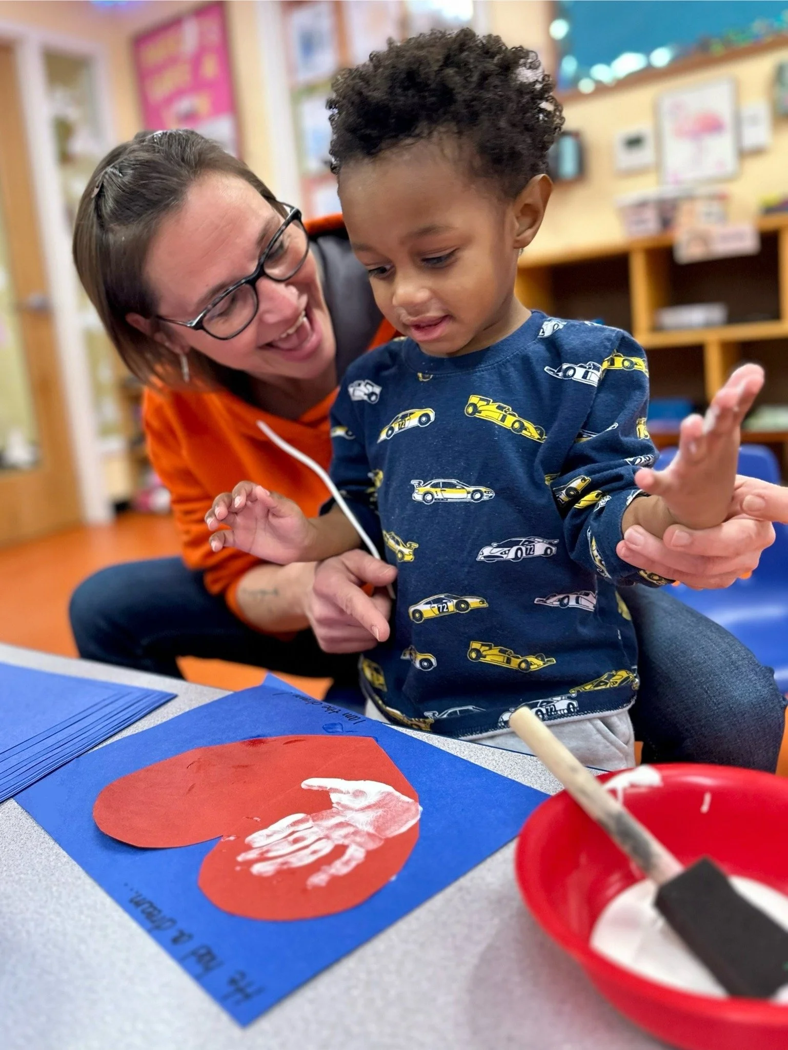 A young child and a woman, likely a teacher or caregiver, are engaging in a craft activity at a classroom table. The child is wearing a blue shirt with yellow cars, and the woman is smiling and assisting. On the table, there is a blue sheet of paper with a red heart-shaped cutout and white handprints, along with a red bowl containing art supplies.