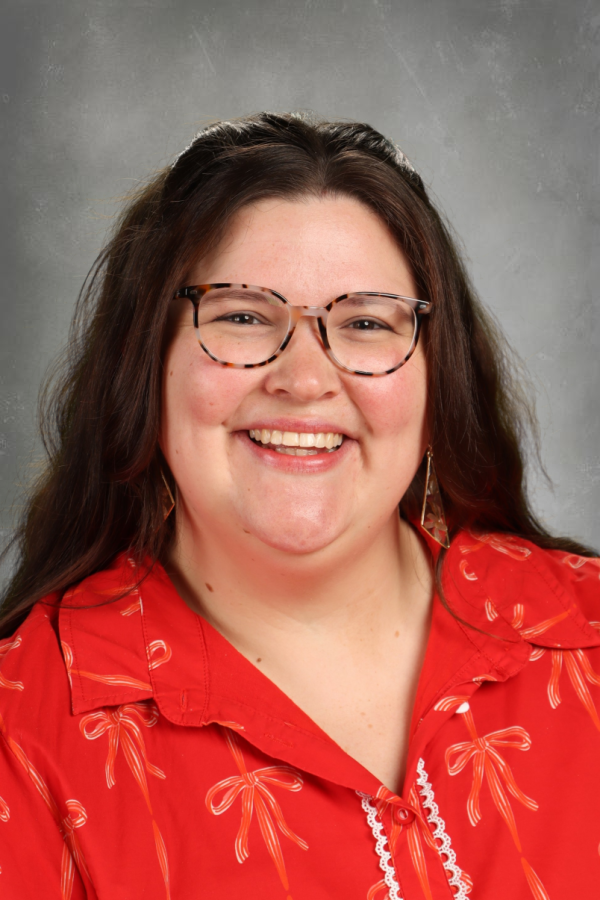 A woman with long brown hair, wearing glasses, a red shirt with white bow designs, earrings, and smiling in front of a gray background.