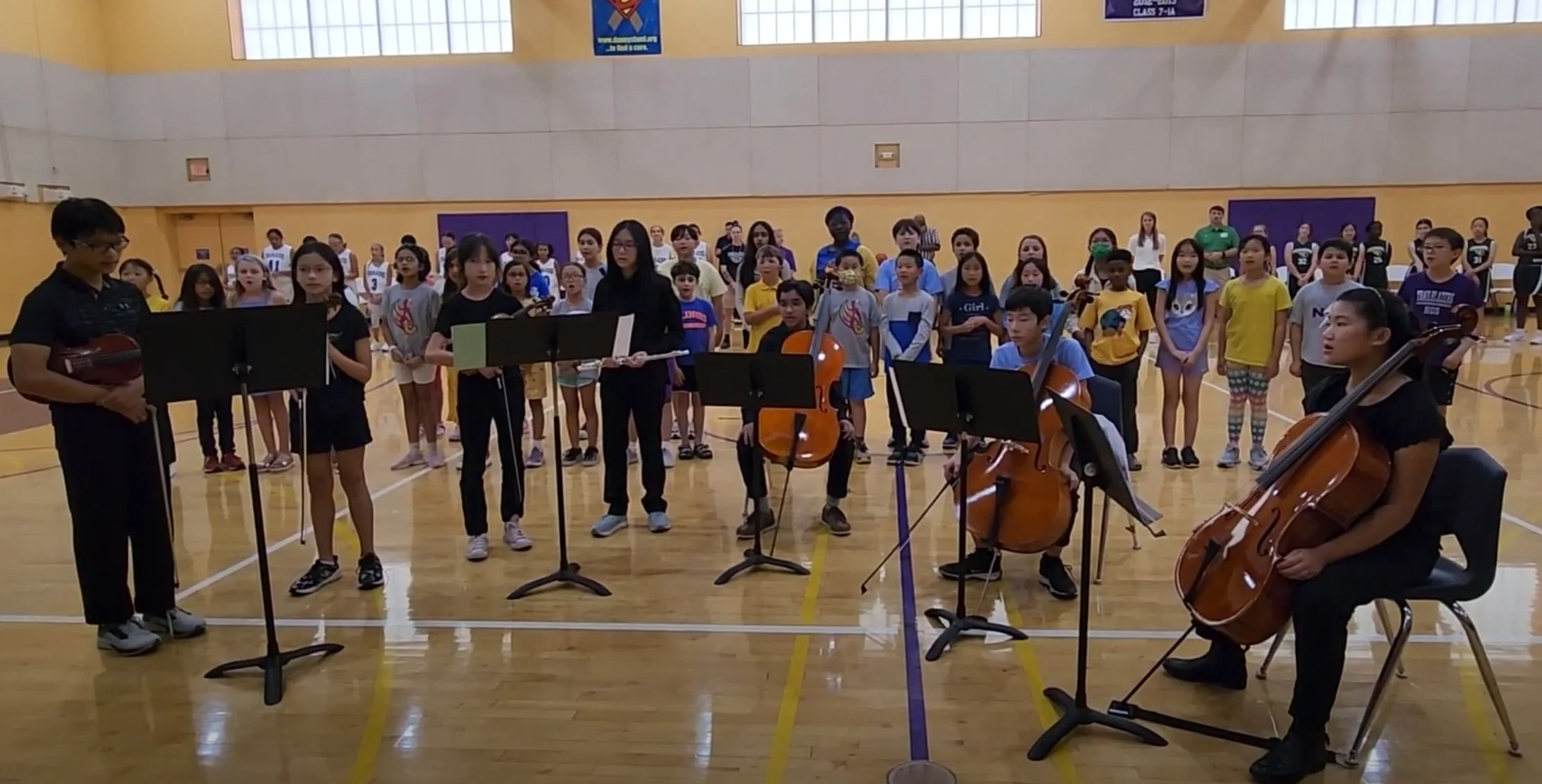 A group of children performing with musical instruments in a gymnasium, with an audience of children standing behind them.