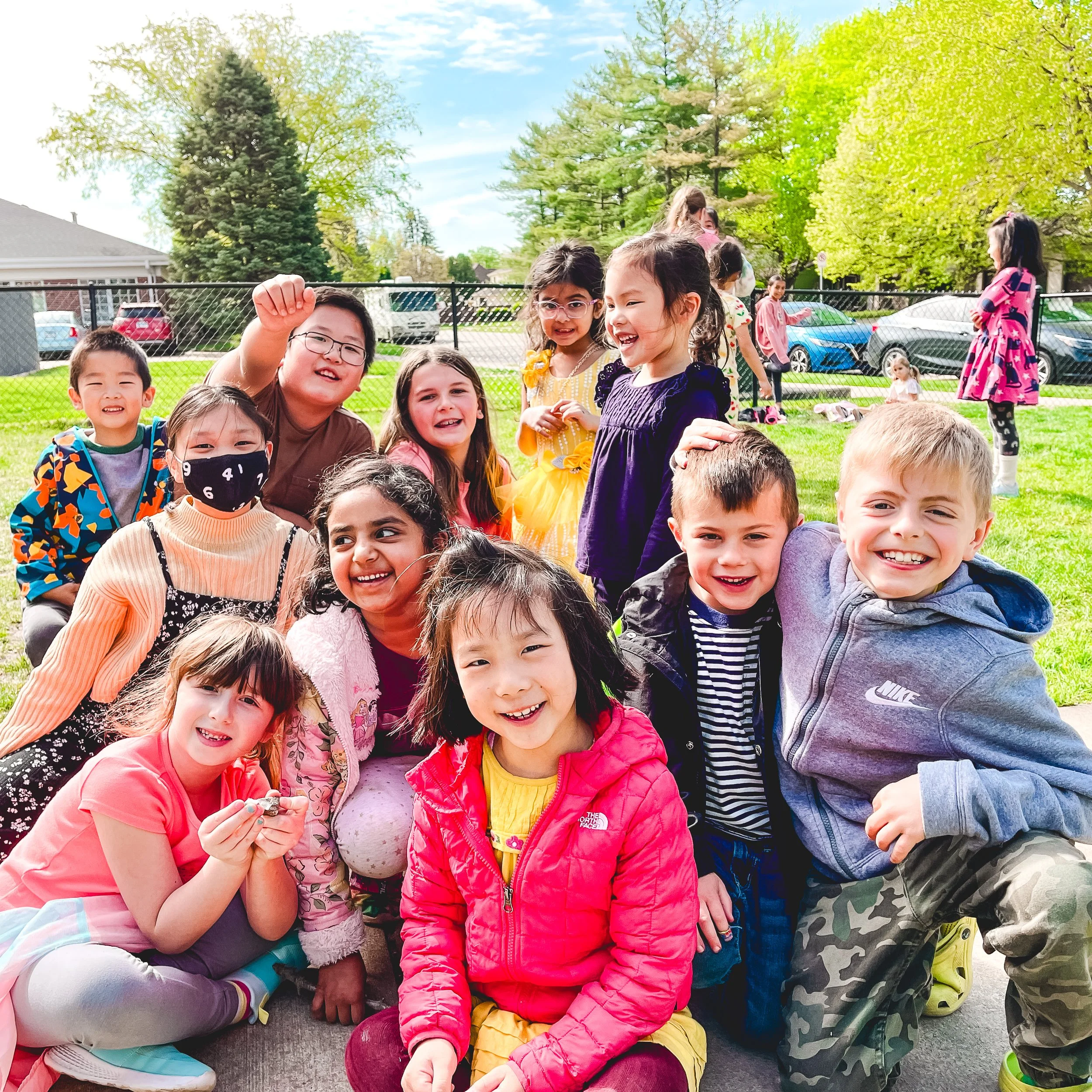 Group of children outdoors on a sunny day, smiling and interacting with each other, with trees, a fence, and parked cars in the background.
