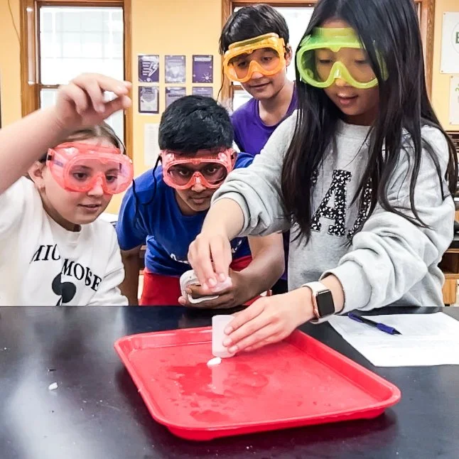 Four children wearing safety goggles watch as a girl in a gray hoodie demonstrates a science experiment with dry ice on a red tray.