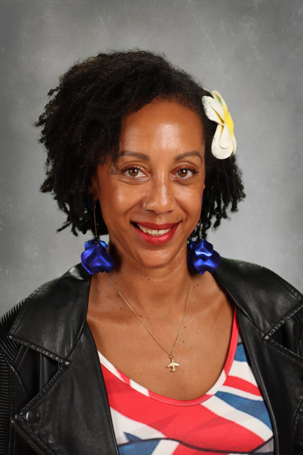 A woman with natural curly hair wearing a white flower hair accessory, blue earrings, a black leather jacket, and a shirt with red, white, and blue patterns, smiling against a gray background.