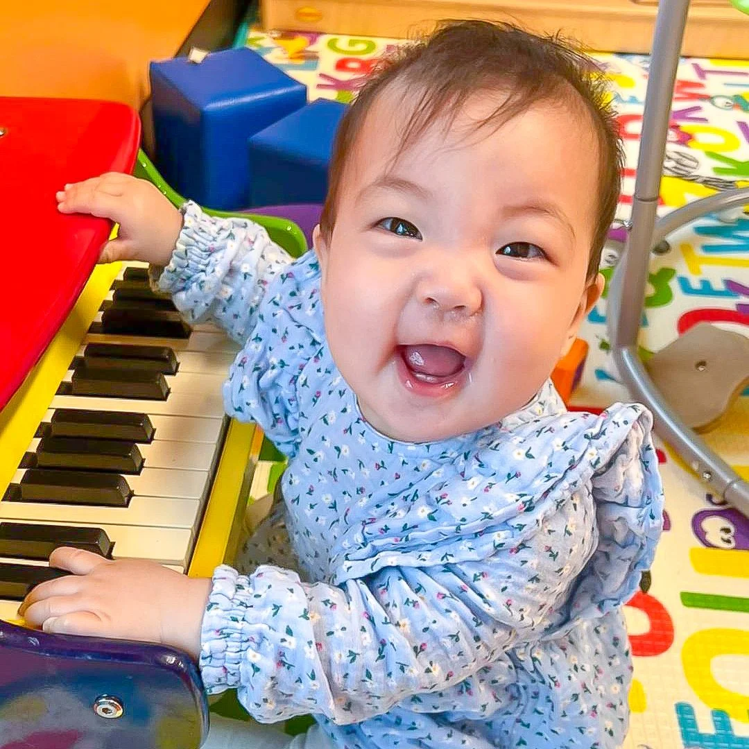 A happy baby with a toothless smile playing on a colorful toy piano in a playroom.
