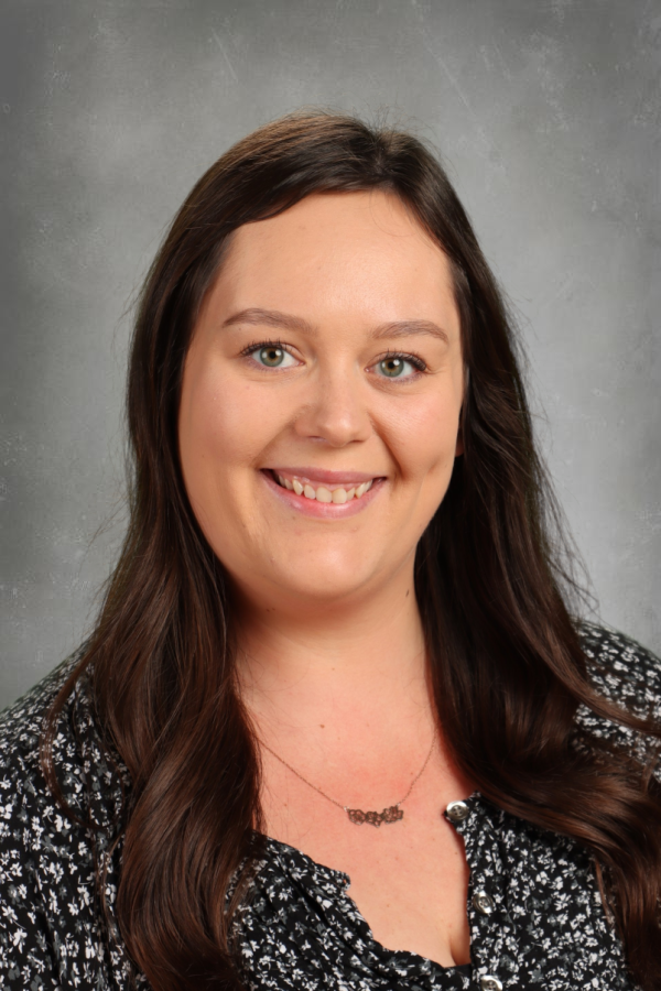 A woman with long brown hair smiling, wearing a black and white patterned blouse and a necklace against a gray background.