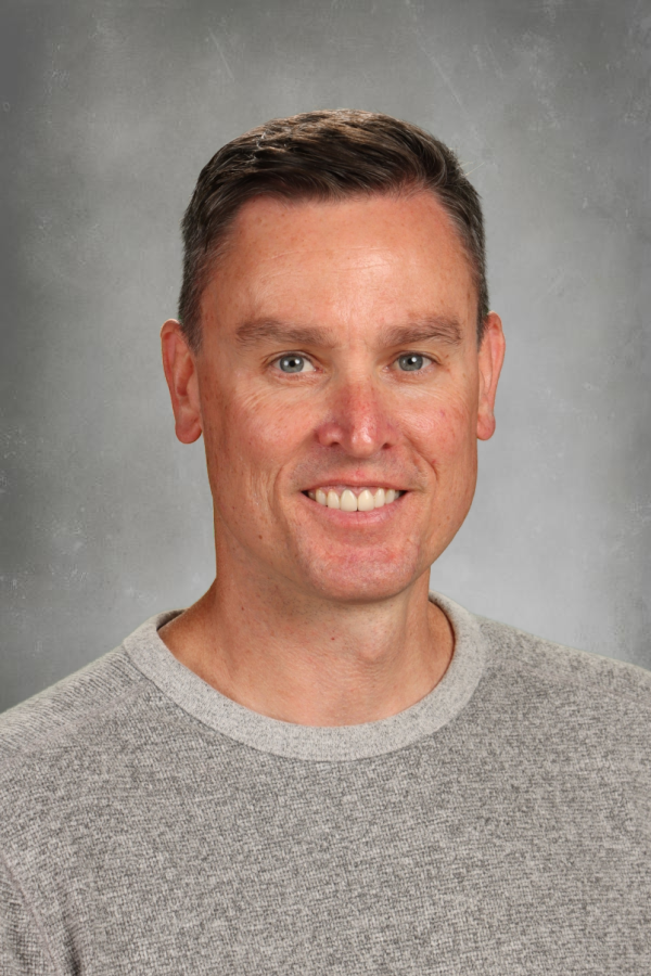 A smiling man with short brown hair and blue eyes, wearing a gray sweater, standing against a gray background.