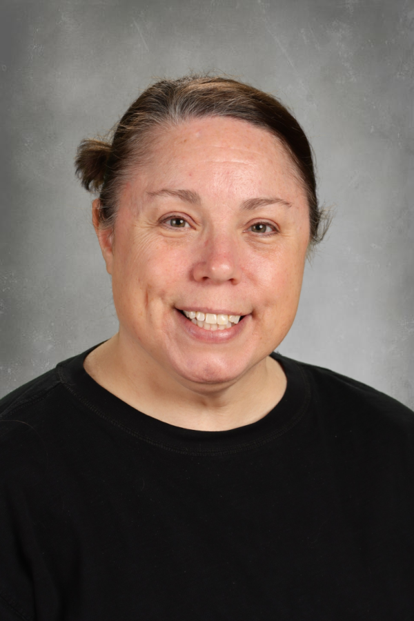 A woman with brown hair in a small bun, smiling slightly, wearing a black shirt, against a gray textured background.
