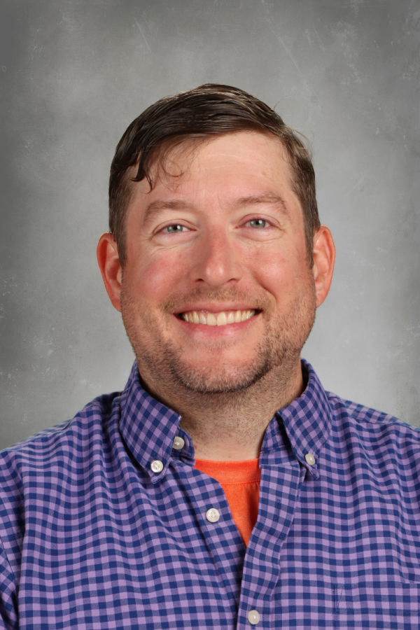 A smiling man with short brown hair and light stubble, wearing a purple checkered button-up shirt over an orange t-shirt, against a gray background.
