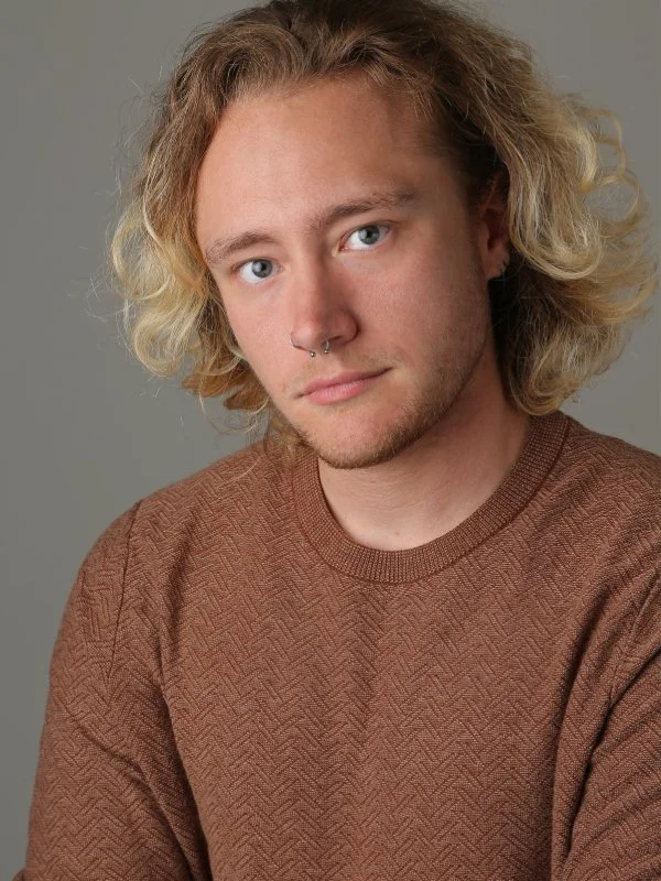 A young man with curly blond hair and blue eyes, wearing a brown textured sweater, looking at the camera against a gray background.