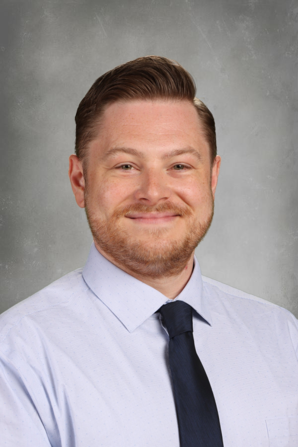 Headshot of a man with styled hair and a beard, smiling, wearing a light blue dress shirt and a dark tie, against a gray background.
