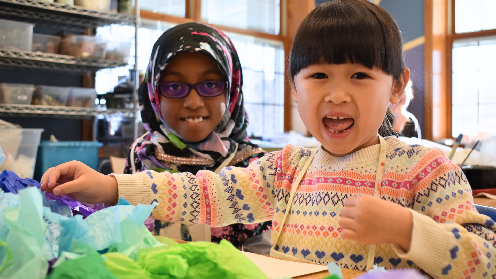 Two happy children at a craft table with tissue paper colorful decorations. The girl in the background wears glasses and a patterned headscarf, the girl in the foreground has short dark hair and is wearing a colorful sweater.