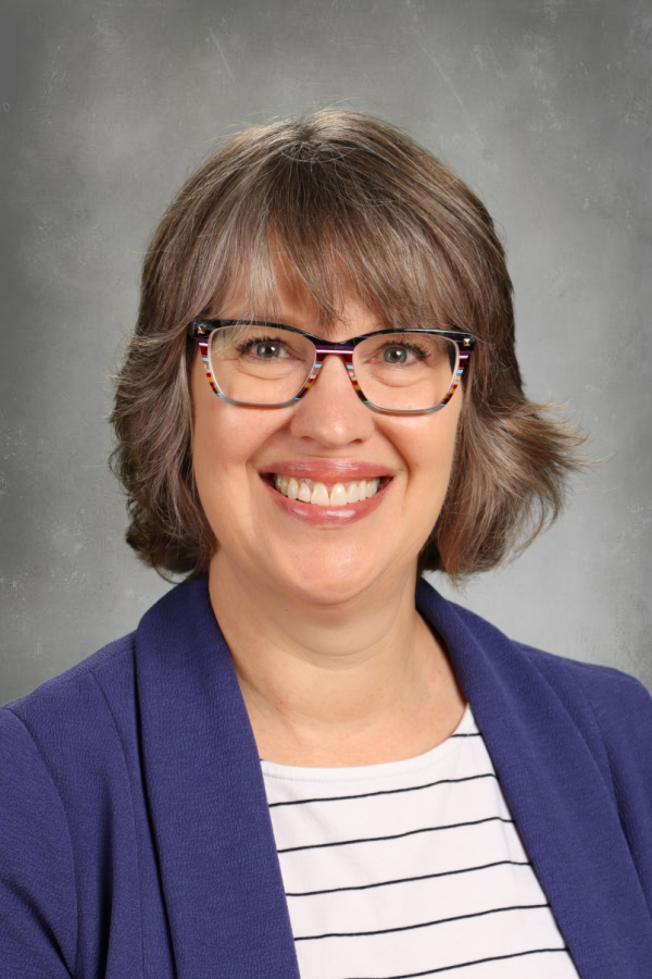 A woman smiling, wearing glasses, a white and black striped shirt, and a blue blazer, with gray hair and a neutral gray background.