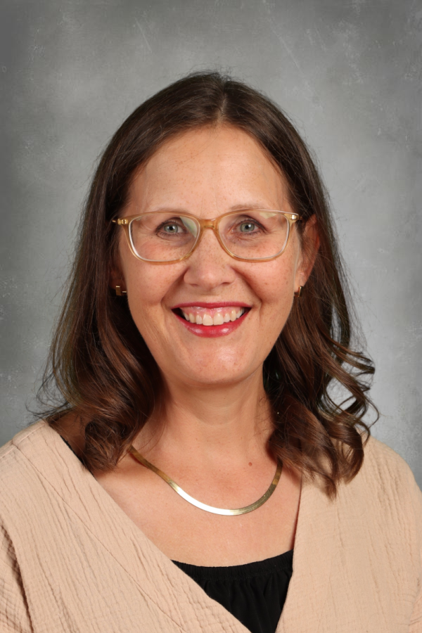 A woman with shoulder-length brown hair, wearing glasses, a beige top, and a gold necklace, smiling against a gray background.