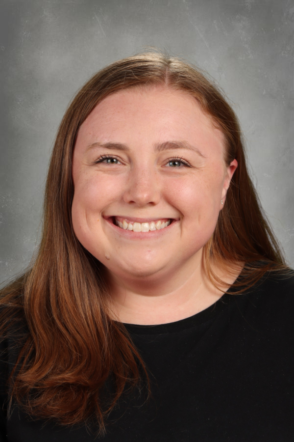 Photograph of a young woman with long brown hair, smiling, wearing a black top, against a gray background.