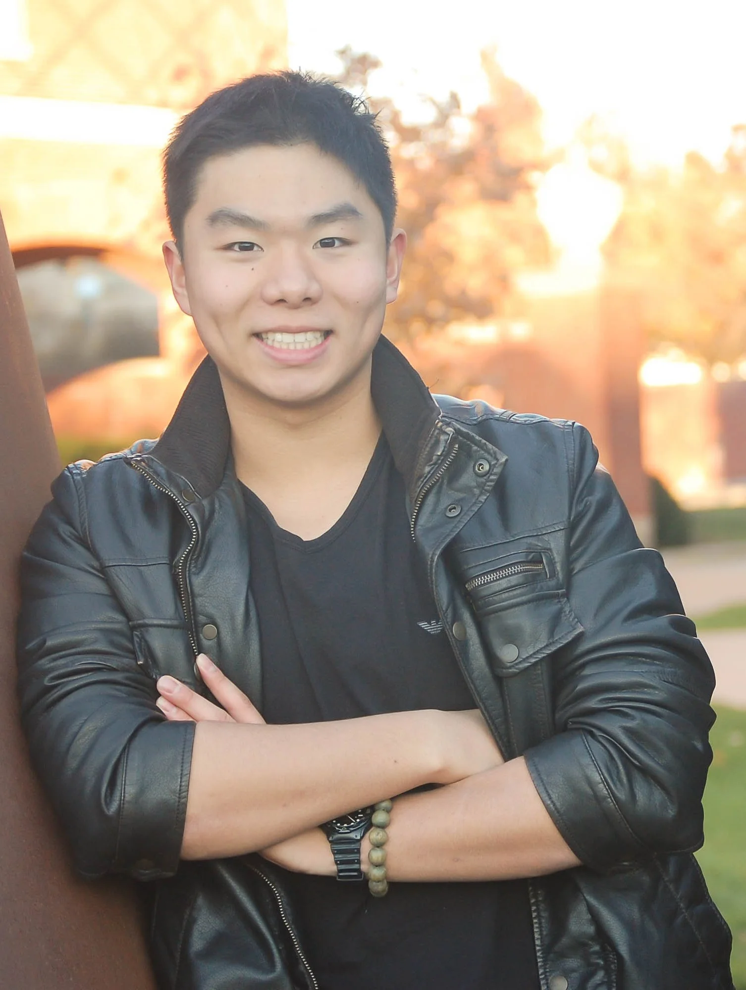 A young man standing outdoors, smiling with arms crossed. He's wearing a black leather jacket, a black t-shirt, and a beaded bracelet, with trees and warm sunlight in the background.
