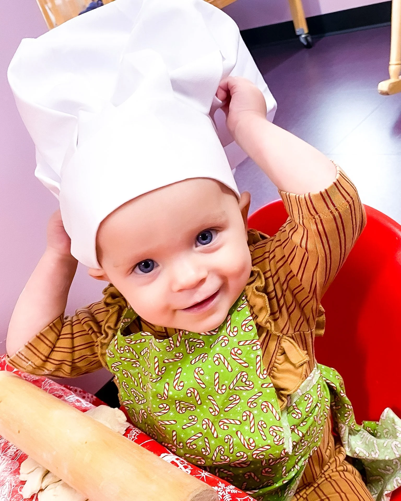 Baby boy wearing a chef hat and a festive apron, sitting at a table with a pizza dough and flour, smiling at the camera.
