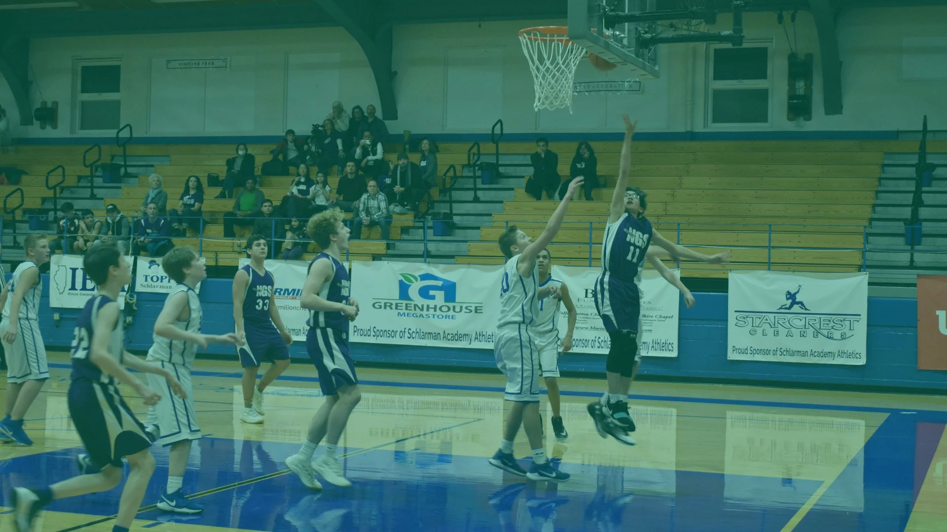 A youth basketball game in progress with players jumping and reaching for the ball near the hoop in a gymnasium with spectators in the background.