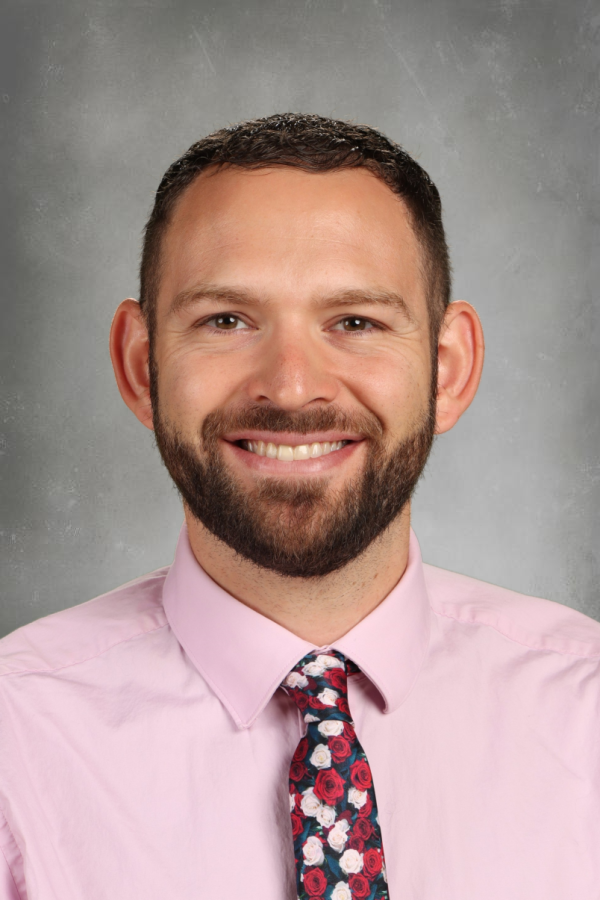Portrait of a smiling man with short dark hair and beard, wearing a light pink dress shirt and a floral tie, against a gray background.