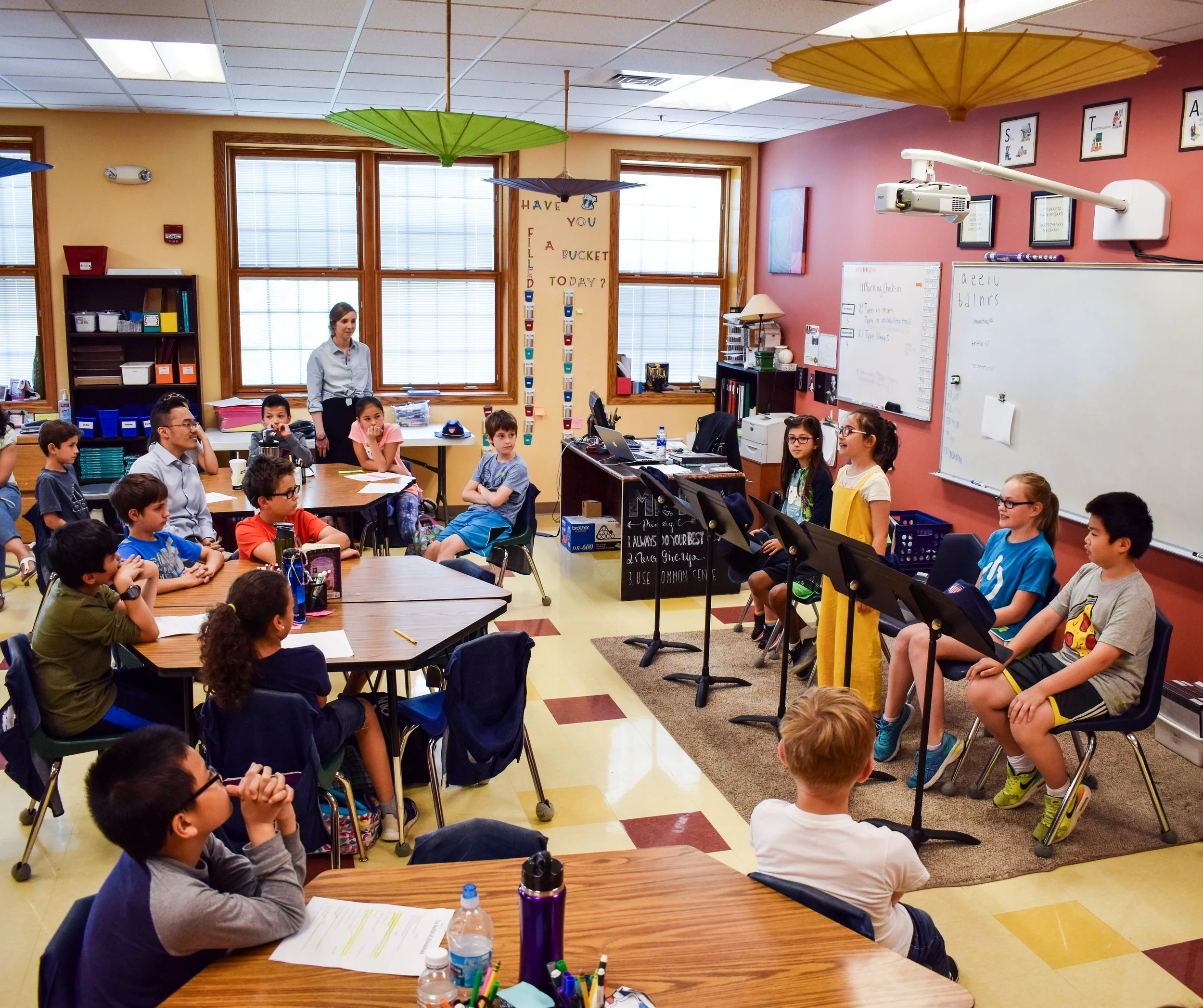 A classroom with students sitting at tables and listening to four students playing recorders at the front. A teacher is standing near the windows, and colorful umbrellas hang from the ceiling.