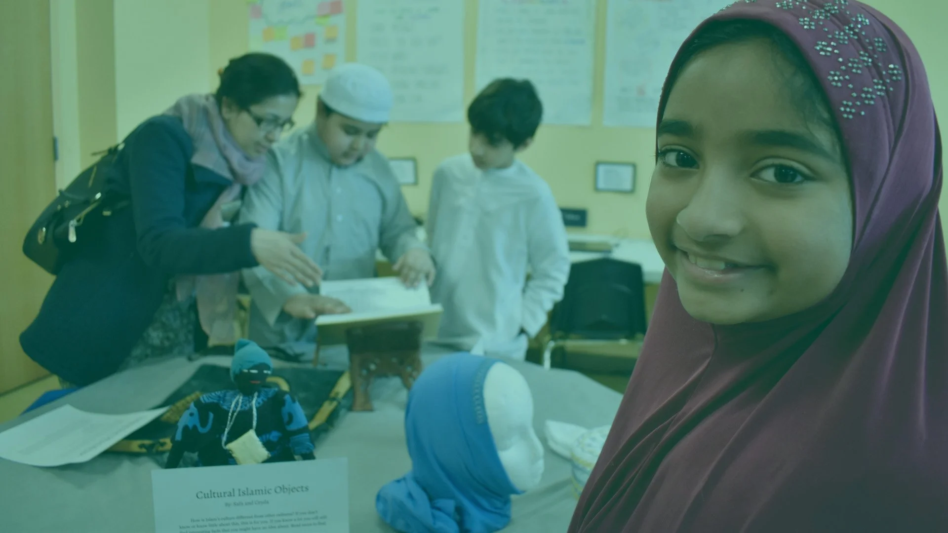 A smiling young girl in a purple hijab stands near display tables with cultural Islamic objects, while in the background, three people are examining items on another table in a classroom or exhibition setting.
