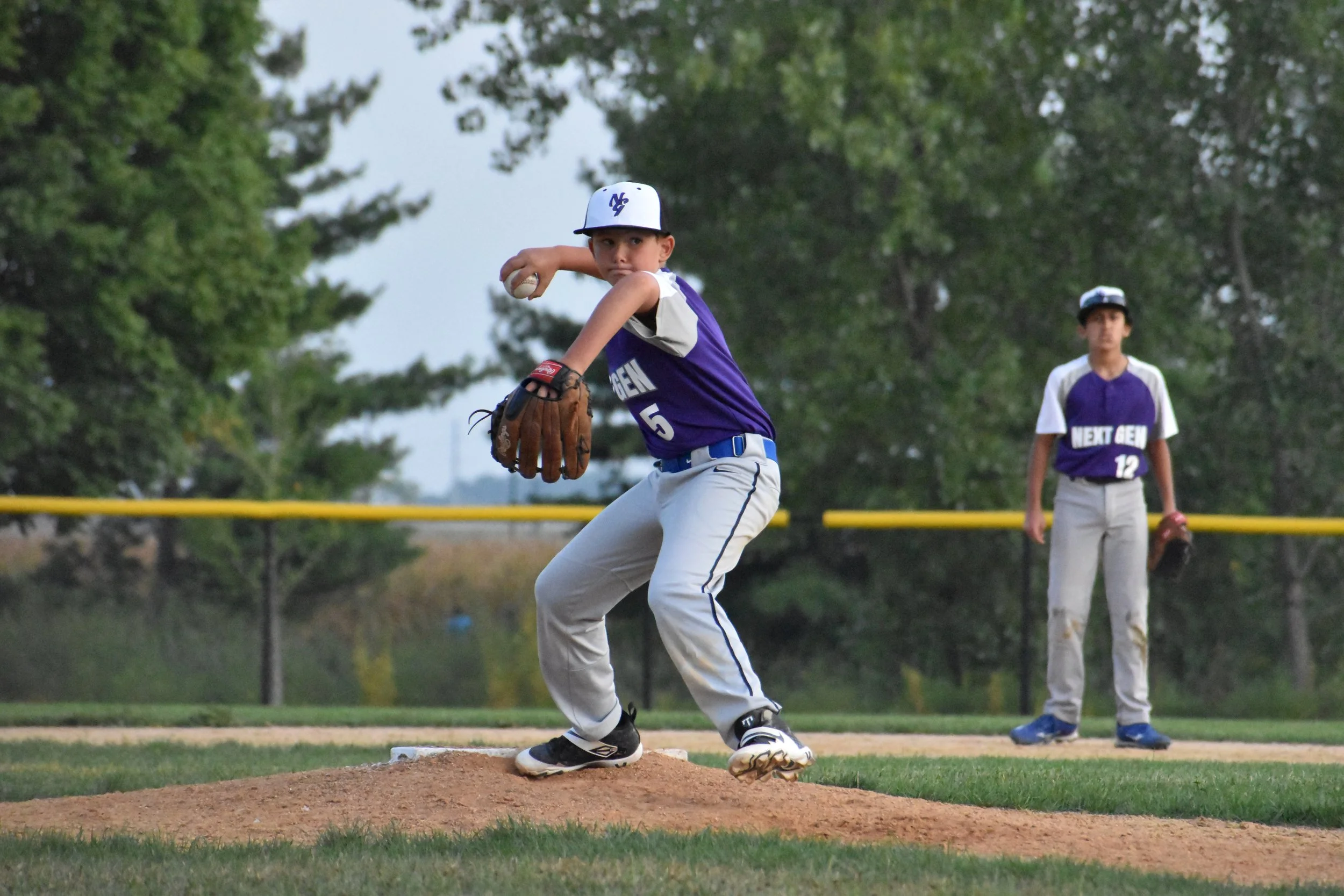 Young baseball player in purple jersey and gray pants crouches on the pitcher's mound, preparing to pitch, with another player in similar uniform standing behind him on the field.
