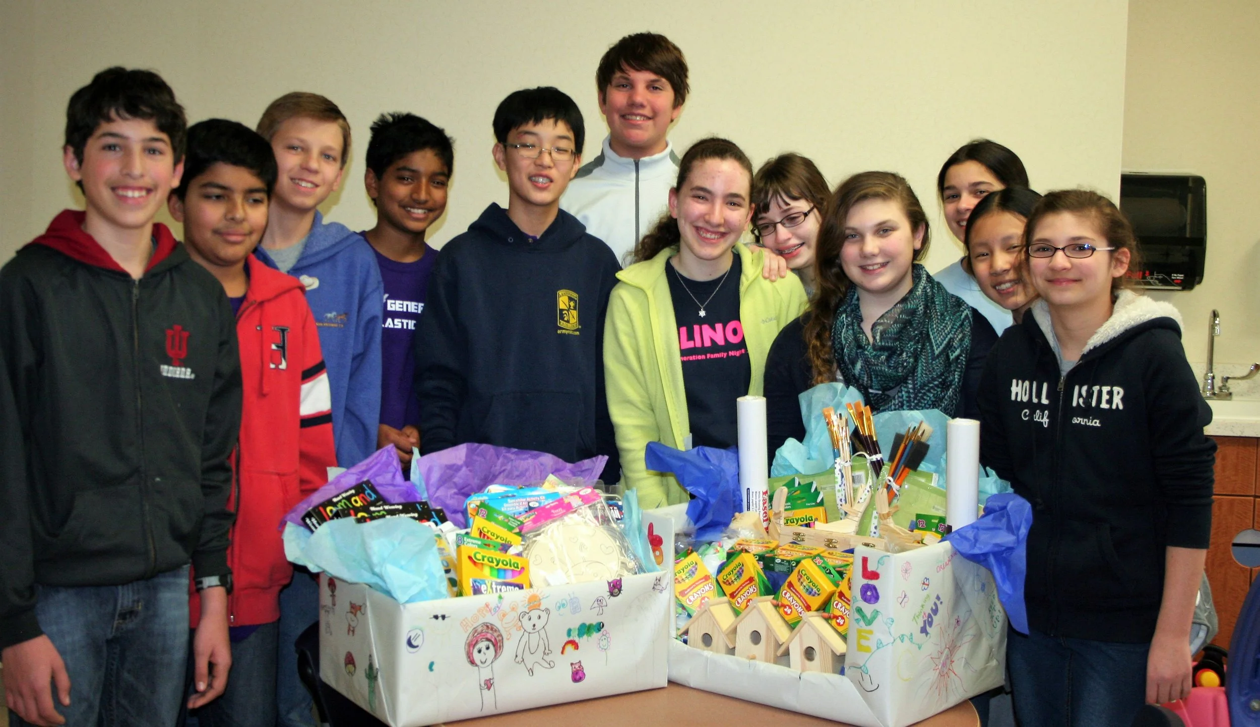 Group of children and teenagers smiling behind a table filled with art supplies and school supplies, in a classroom setting.