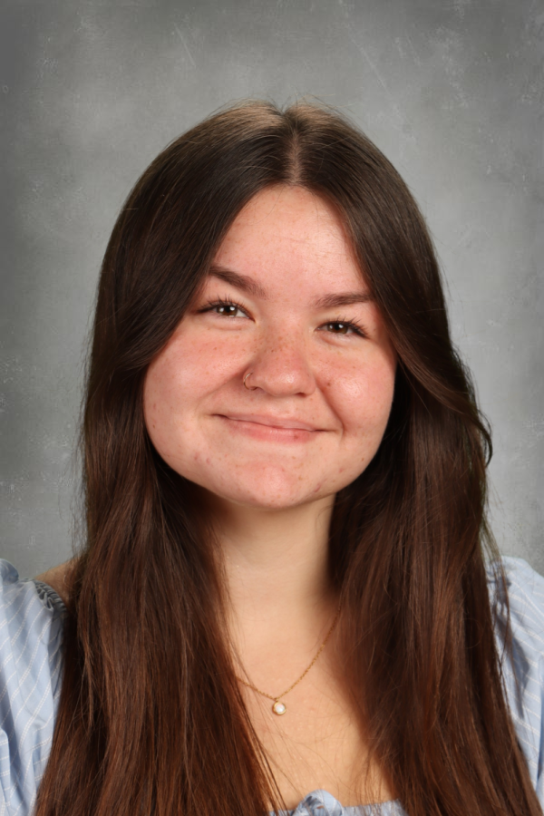 A young woman with long brown hair smiling, wearing a light blue top and a gold necklace, against a gray background.
