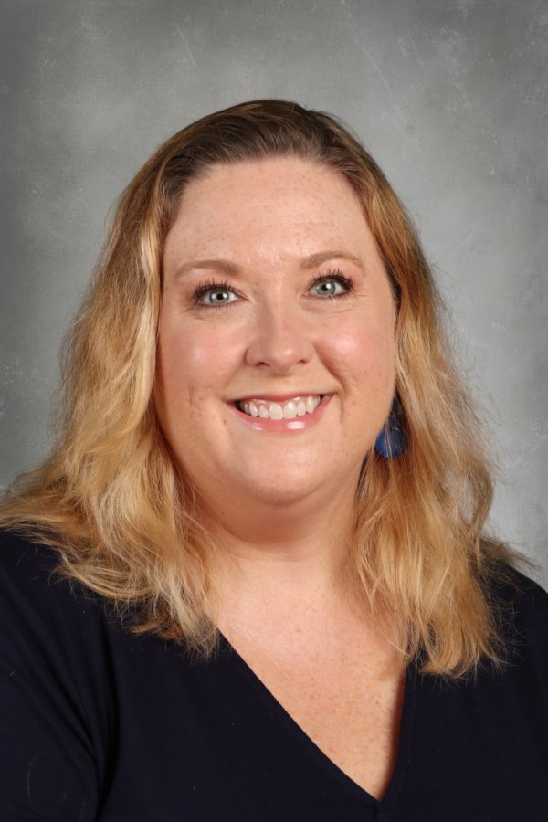 A woman with shoulder-length wavy blonde hair, blue eyes, and light skin, smiling in front of a gray background, wearing a black top and blue earrings.