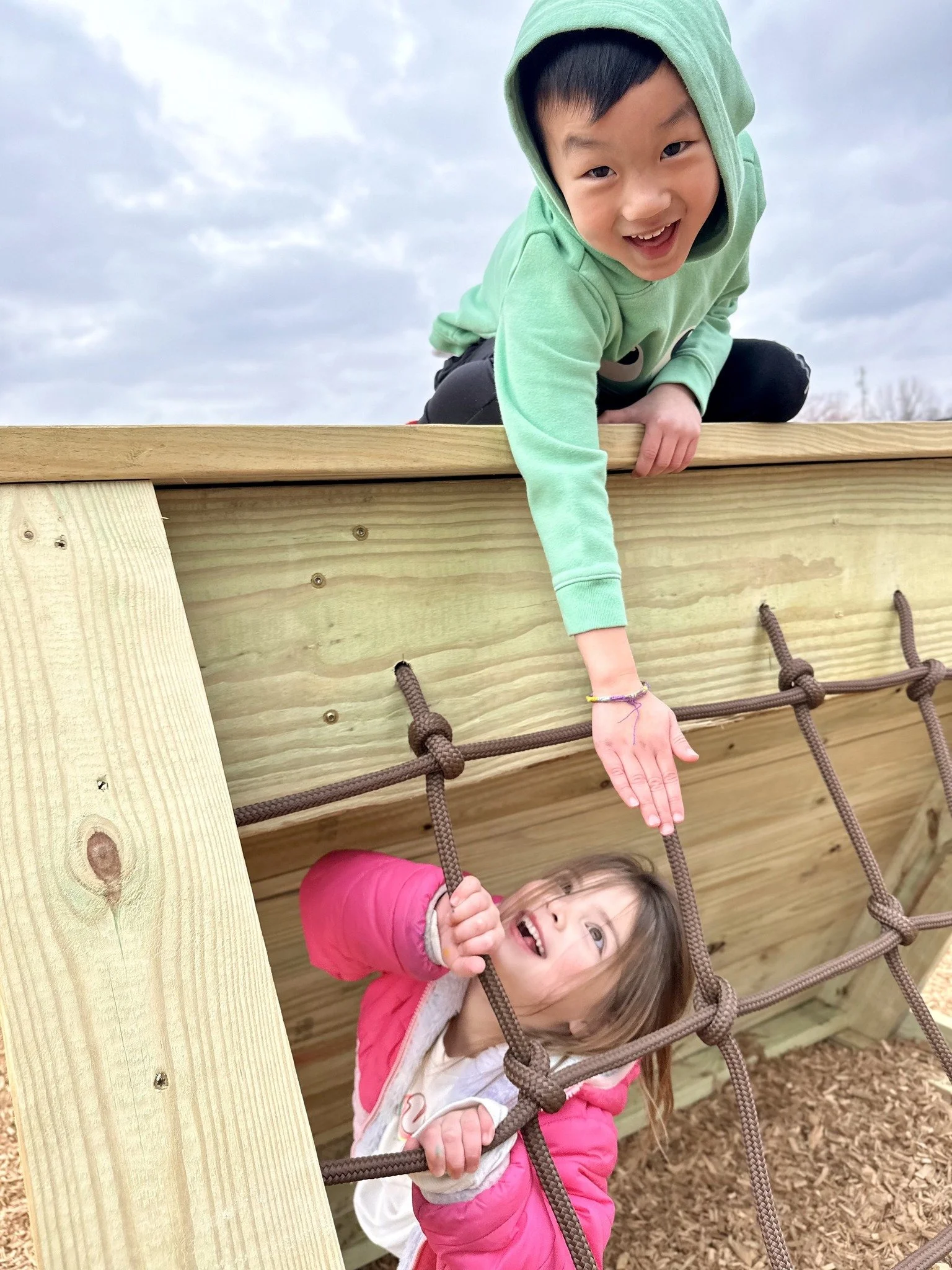 Two children playing on a wooden playground structure, with one child reaching down to help the other up the rope ladder, outdoors on a cloudy day.