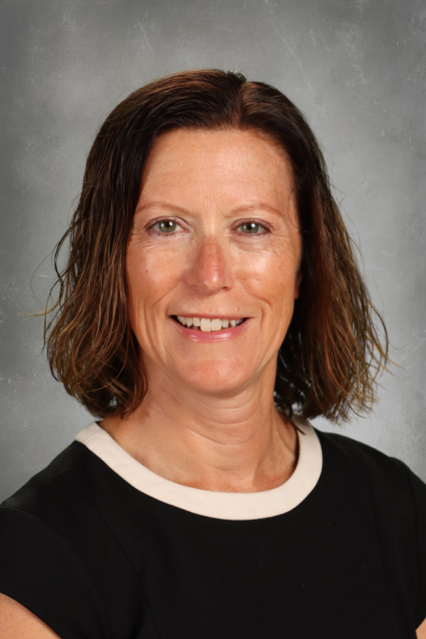 A woman with shoulder-length wavy brown hair, smiling, wearing a black shirt with a white collar, against a gray textured background.