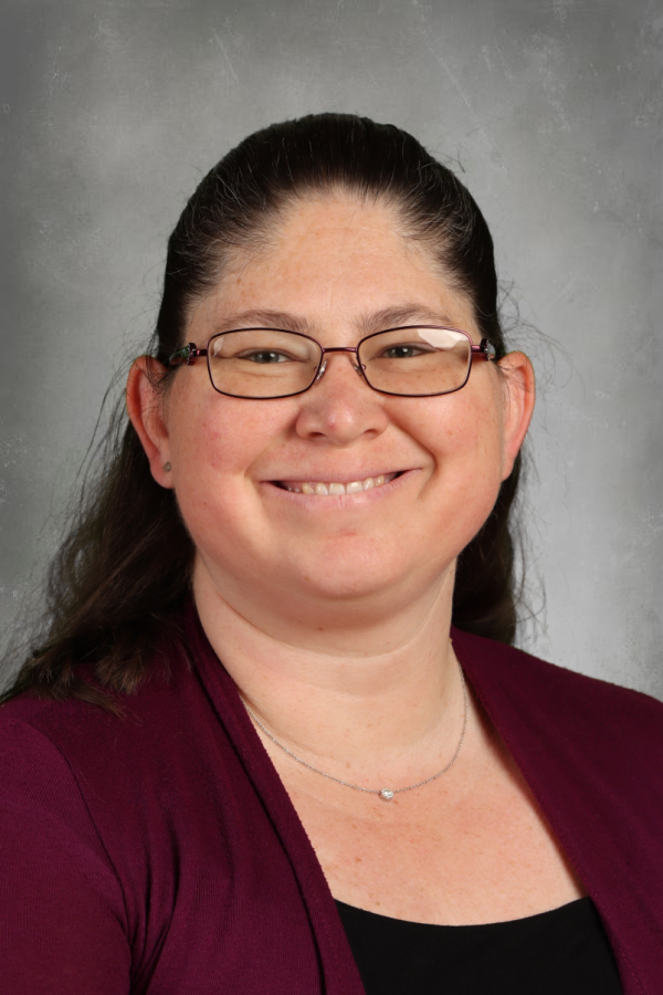 A woman with glasses, smiling, with dark hair pulled back, wearing a burgundy jacket and a black top, against a gray background.