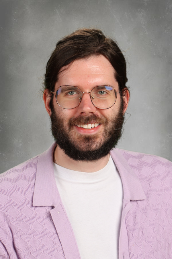 Portrait of a man with glasses, a beard, wearing a white shirt and a light purple patterned jacket, smiling against a neutral background.