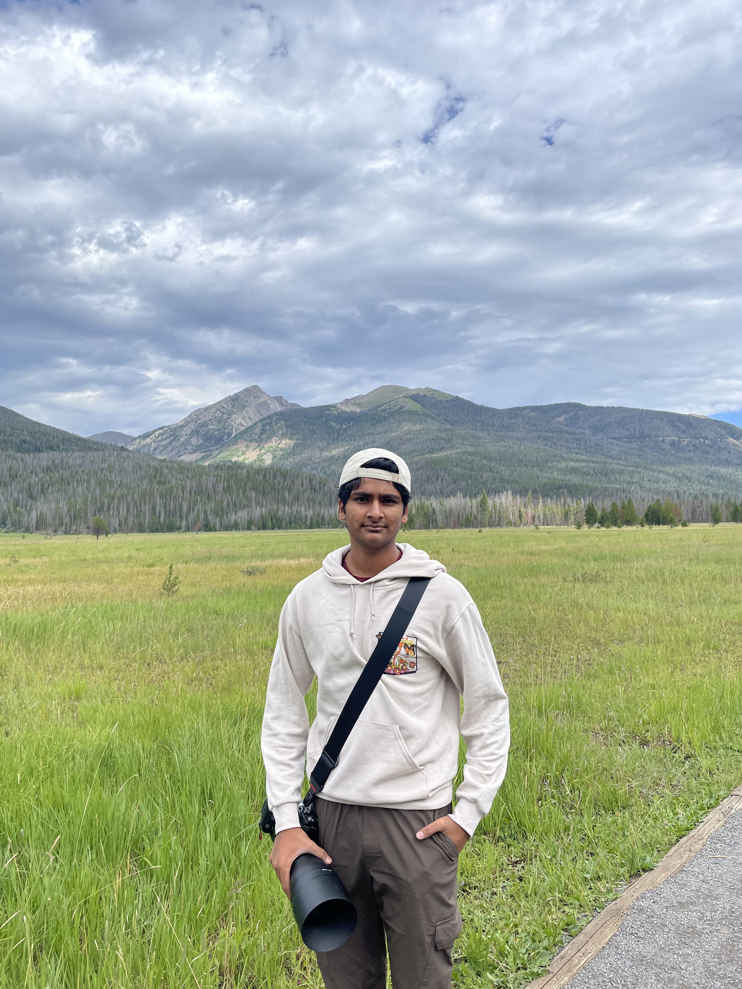 A young man standing outdoors in a grassy field with mountains and cloudy sky in the background. He is wearing a white hoodie, brown pants, a backward white cap, and holding a camera with a large lens.